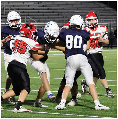 People wearing football uniforms are engaged in a play on a green field with empty bleachers in the background. Description generated by AI