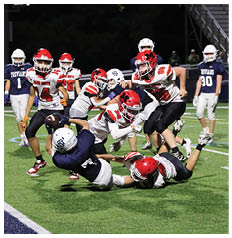 A group of people wearing football uniforms are actively engaged in a tackle on a well-lit field with a few others standing in the background near bleachers. Description generated by AI