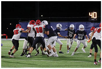 Players in red and white helmets and navy and white helmets are engaged in a football game on a green field under stadium lights. Description generated by AI