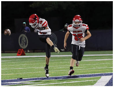 Two people in football uniforms with red helmets are on a field, with one kicking a football while the other runs nearby. Description generated by AI