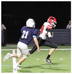 A person in a red and white football uniform is running with the ball while a person in a blue and white uniform and a referee in a black and white striped shirt watch on a football field at night. Description generated by AI