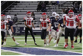 People in football uniforms are actively playing on a field with a football on the ground near the center, while spectators sit sparsely in the bleachers behind them. Description generated by AI