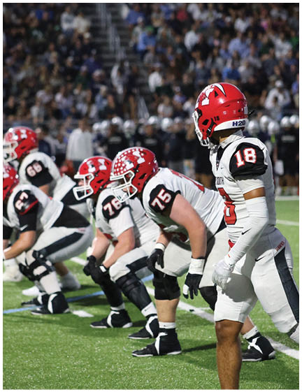 Five football players in white and black uniforms with red helmets are lined up on a green field, preparing for a play. Description generated by AI