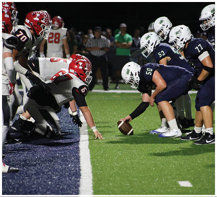 Two people in football uniforms with red helmets are engaged in a play on a green field with others in dark uniforms standing in the background. Description generated by AI
