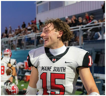 A person wearing a Maine South football jersey is smiling and looking to the side with a blurred crowd in the background. Description generated by AI