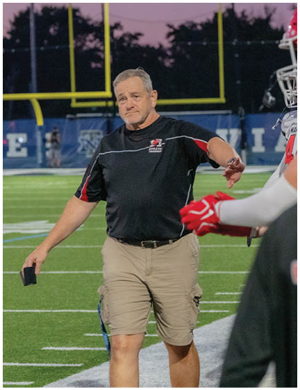 People wearing football helmets and uniforms are standing on a field while another person in a black shirt with a red and white logo is in the foreground. Description generated by AI