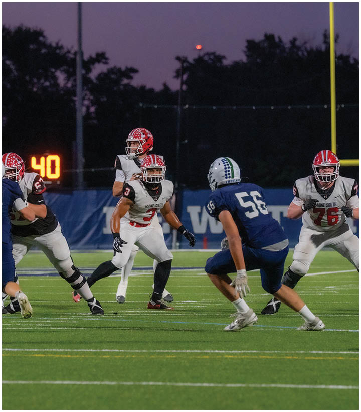 Four people in football uniforms are on a field at dusk with one person in a blue uniform facing three people in white uniforms with red helmets. Description generated by AI