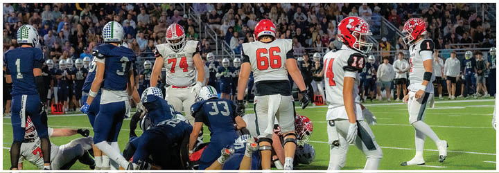 Several people in sports uniforms are gathered on a grassy field with some standing and others on the ground, suggesting a moment during a football game. Description generated by AI