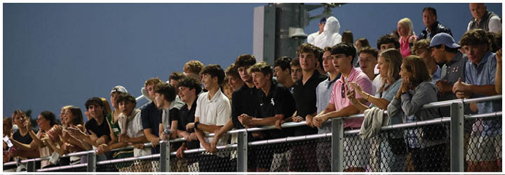 A group of people standing closely together behind a chain-link fence, watching an event with expressions of interest and engagement. Description generated by AI