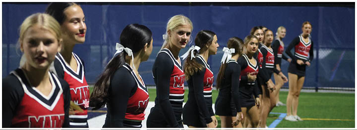 A line of people wearing matching cheerleading uniforms with red and black colors stand on a sports field at night. Description generated by AI