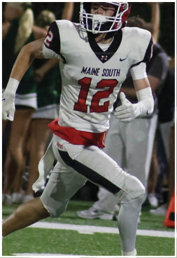 A person wearing a red and white football helmet with the word “HAWKS" on it is looking towards the camera with another person in football gear blurred in the background. Description generated by AI