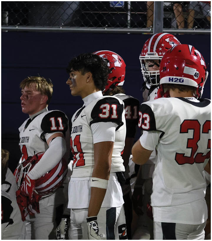 People wearing white and red football uniforms stand together on the sidelines during a game. Description generated by AI