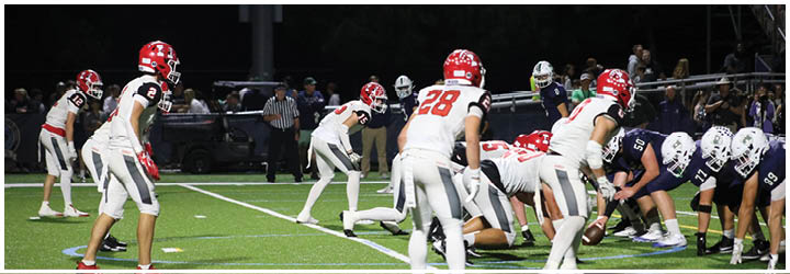 People in football uniforms are lined up on a field at night preparing for a play during a game. Description generated by AI
