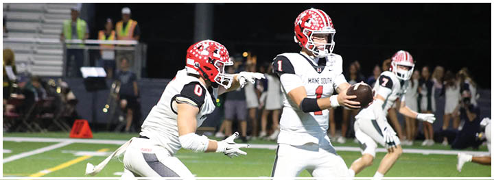 Two people in football uniforms with red helmets are on a field at night, with one holding a football and the other reaching out toward them. Description generated by AI