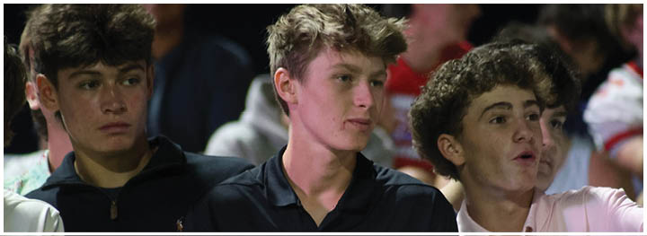 Three young people stand close together, with one wearing a black sports jacket, another in a black polo shirt with a white logo, and the third in a light-colored shirt, all appearing engaged in conversation or observation. Description generated by AI