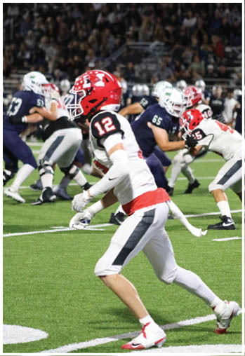 A person wearing a red helmet and white football uniform with the number 12 is running on a football field while other players in dark and white uniforms are engaged in the background. Description generated by AI