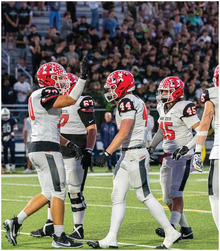 Five people wearing white and gray football uniforms with red helmets are standing on a football field with a crowd in the background. Description generated by AI