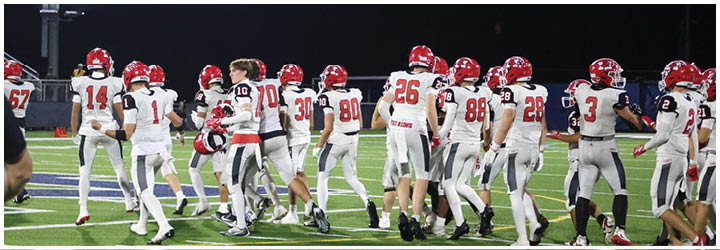 A group of people wearing white and red football uniforms are gathered on a green football field at night with a person in black standing in the foreground. Description generated by AI