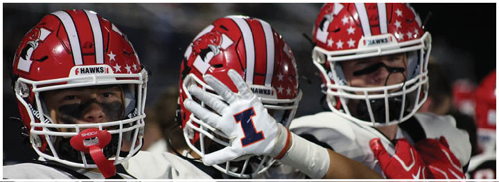 Three people wearing red and white football helmets with a hawk logo and black face paint are standing close together. Description generated by AI