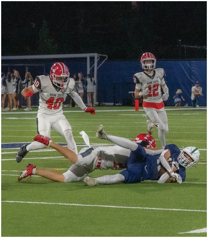Two people in football uniforms are tackling a third person holding a football on a field while a referee and other players watch in the background. Description generated by AI