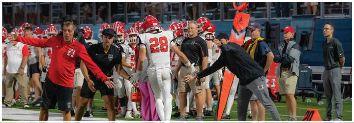 People in sports uniforms and coaches are actively engaged on the sidelines of a football field during a game, with a crowd visible in the stands behind them. Description generated by AI