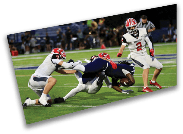 Three people in white sports uniforms tackle a person in a dark blue uniform holding a football on a green field while another person in white watches nearby. Description generated by AI