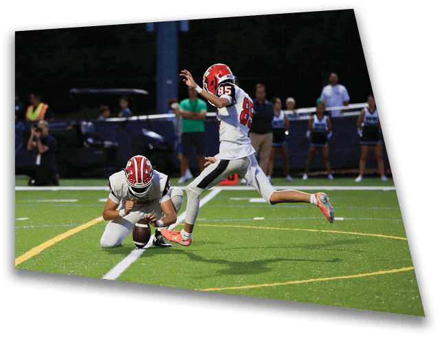 A person in a red helmet is kicking a football held by another person kneeling on a green field during a game with spectators in the background. Description generated by AI