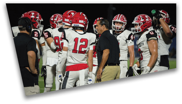 A group of football players in white uniforms with red helmets and a coach in black shirt and khaki pants are gathered on the field during a game. Description generated by AI