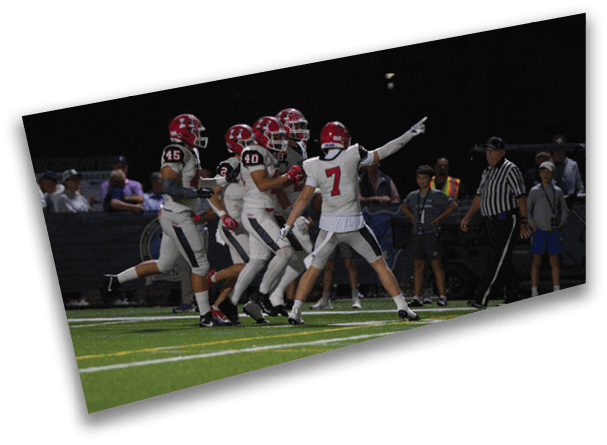 A group of football players in red helmets and white uniforms are celebrating on a field at night while a referee and spectators watch from the sidelines. Description generated by AI