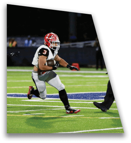 A person wearing a red helmet and football uniform is running with a football on a green field while a referee in a black and white striped shirt walks nearby. Description generated by AI