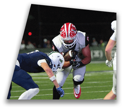 Two people in football uniforms are engaged in a play on a field at night, with one holding the ball and the other attempting to tackle. Description generated by AI