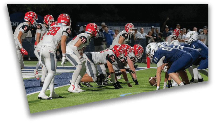 Two opposing American football teams in red and blue uniforms are lined up facing each other on a field, preparing for a play. Description generated by AI