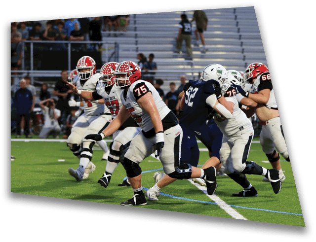 Players in football uniforms are actively running on a green field during a game with spectators in the background. Description generated by AI