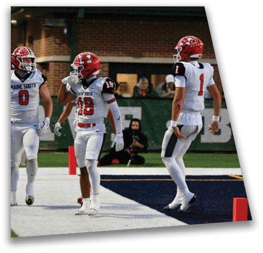 Two people in white football uniforms and red helmets stand on a field with a green banner and spectators in the background. Description generated by AI