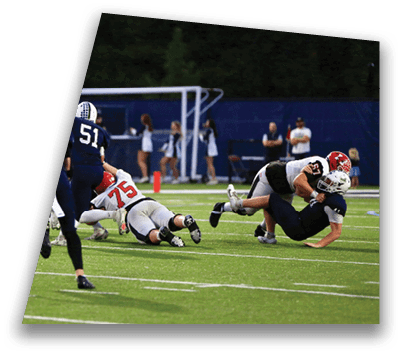 People in football uniforms are actively playing on a field with some players running and others tackling. Description generated by AI