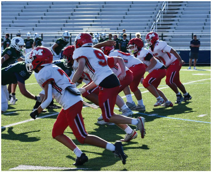Children in red and white football uniforms are lined up on a grassy field preparing to start a play. Description generated by AI