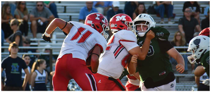Two people in red and white football uniforms are tackling a person in a dark green uniform during a game with spectators in the background. Description generated by AI