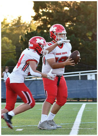 Two people in red and white football uniforms are on a field with one holding a football preparing to throw while the other runs beside them. Description generated by AI