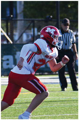 A person in a red and white football uniform is running on a green field with a referee standing in the background. Description generated by AI
