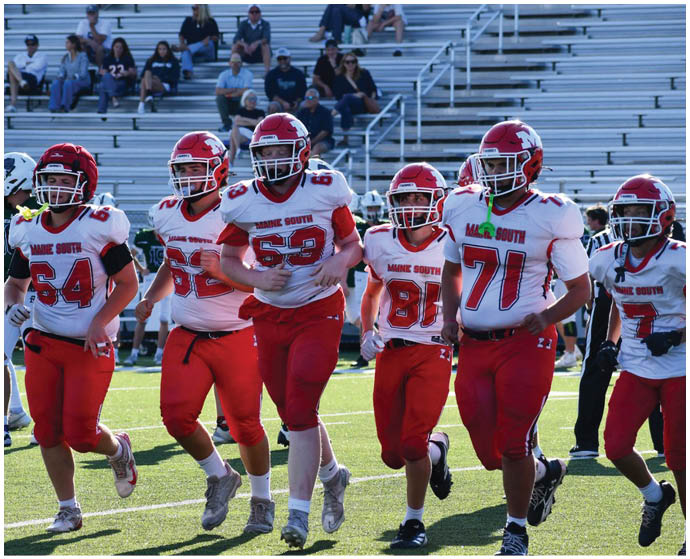 Five people in red and white football uniforms with helmets are walking together on a football field with spectators in the background. Description generated by AI