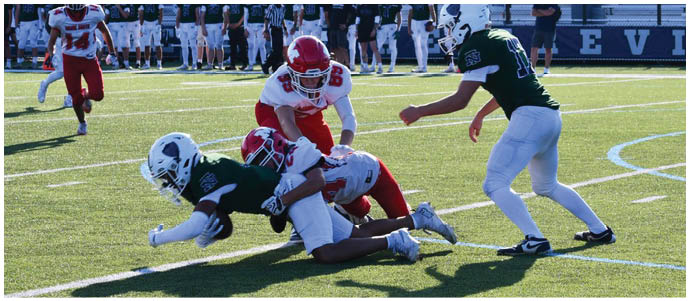 Two people in red and white football uniforms tackle a person in a dark green and white uniform on a football field while another person in a dark green and white uniform approaches. Description generated by AI
