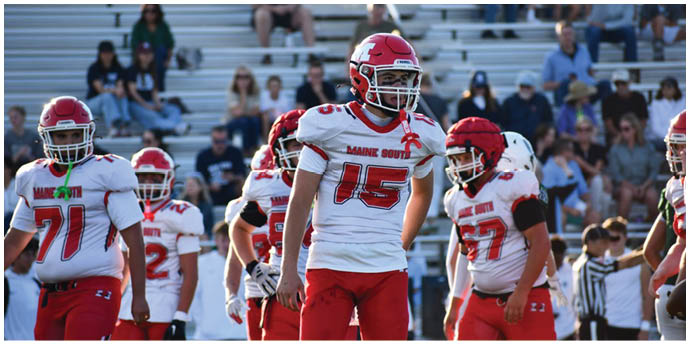 Several people wearing red and white football uniforms with helmets are standing on a field with bleachers and spectators in the background. Description generated by AI