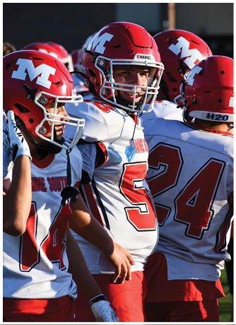 A person wearing a red football helmet with a white “M" and a white jersey is smiling while standing among teammates. Description generated by AI