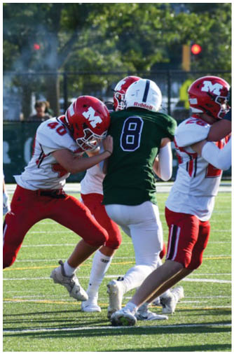 A person in a red and white football uniform is holding a football and extending an arm toward a person in a green and white football uniform running on a field with trees and a scoreboard in the background. Description generated by AI