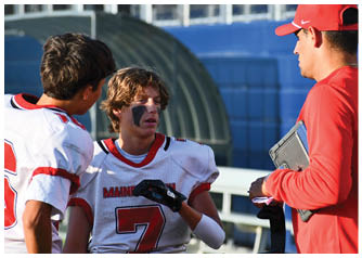 Two people in football uniforms are talking to a third person wearing a red cap and holding a tablet near a sports field. Description generated by AI