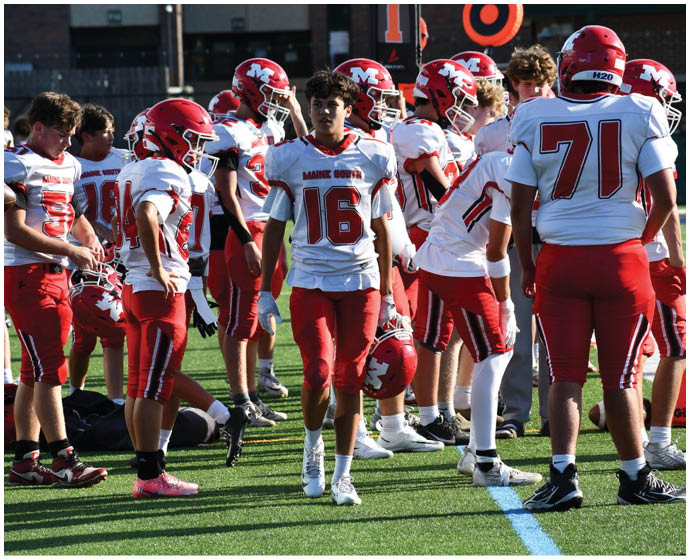 A group of people wearing red and white football uniforms are gathered on a field, some standing and some holding helmets, with one person walking toward the camera. Description generated by AI