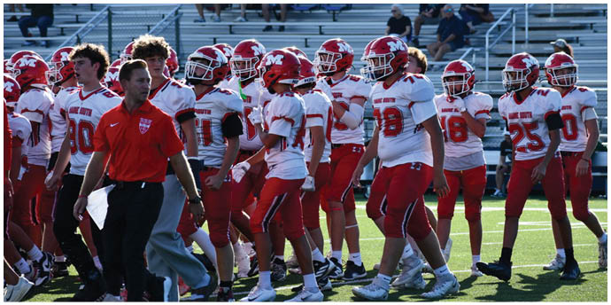 A group of people wearing red and white football uniforms and helmets are gathered on a field with a person in a red shirt walking nearby. Description generated by AI