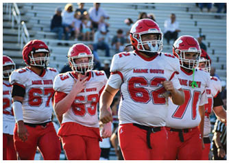 Three people in red and white football uniforms with helmets are on a field, with one person making a hand gesture. Description generated by AI