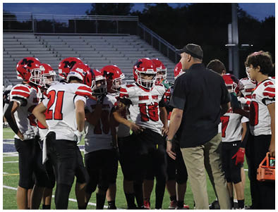 A group of people wearing red and white football uniforms are gathered on a field while a person in casual clothing appears to be giving instructions. Description generated by AI