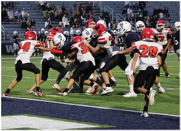 Several people wearing football helmets and uniforms are engaged in a play on a grassy field with a chain marker visible in the background. Description generated by AI